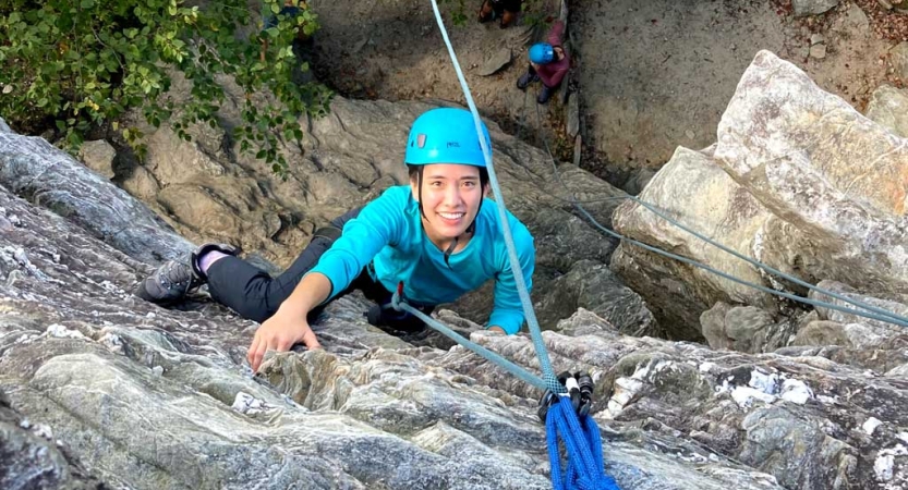 A person wearing safety gear is secured by ropes as they look up at the camera and smile while rock climbing.